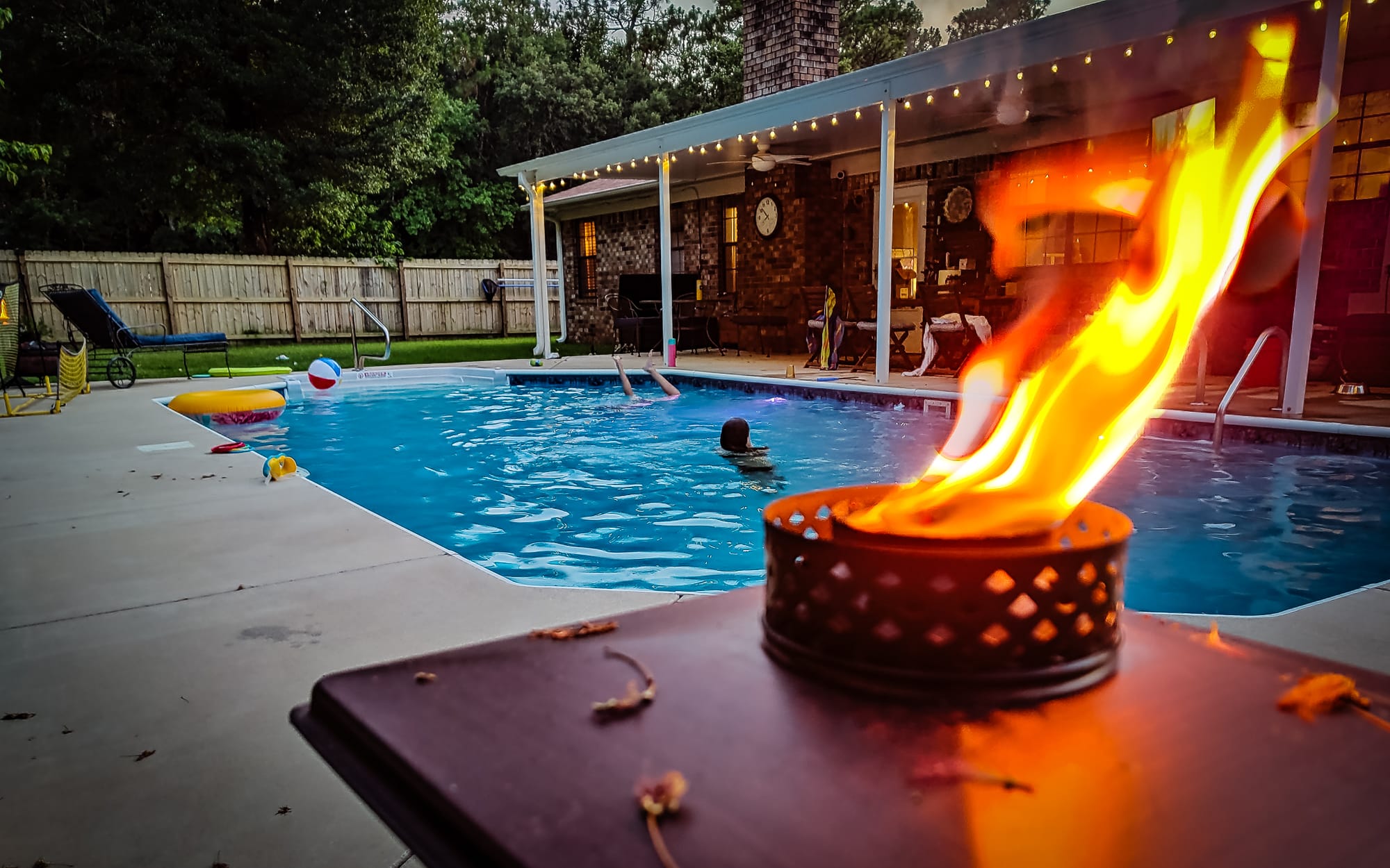 A citronella torch in the foreground as a family swims in a backyard pool.