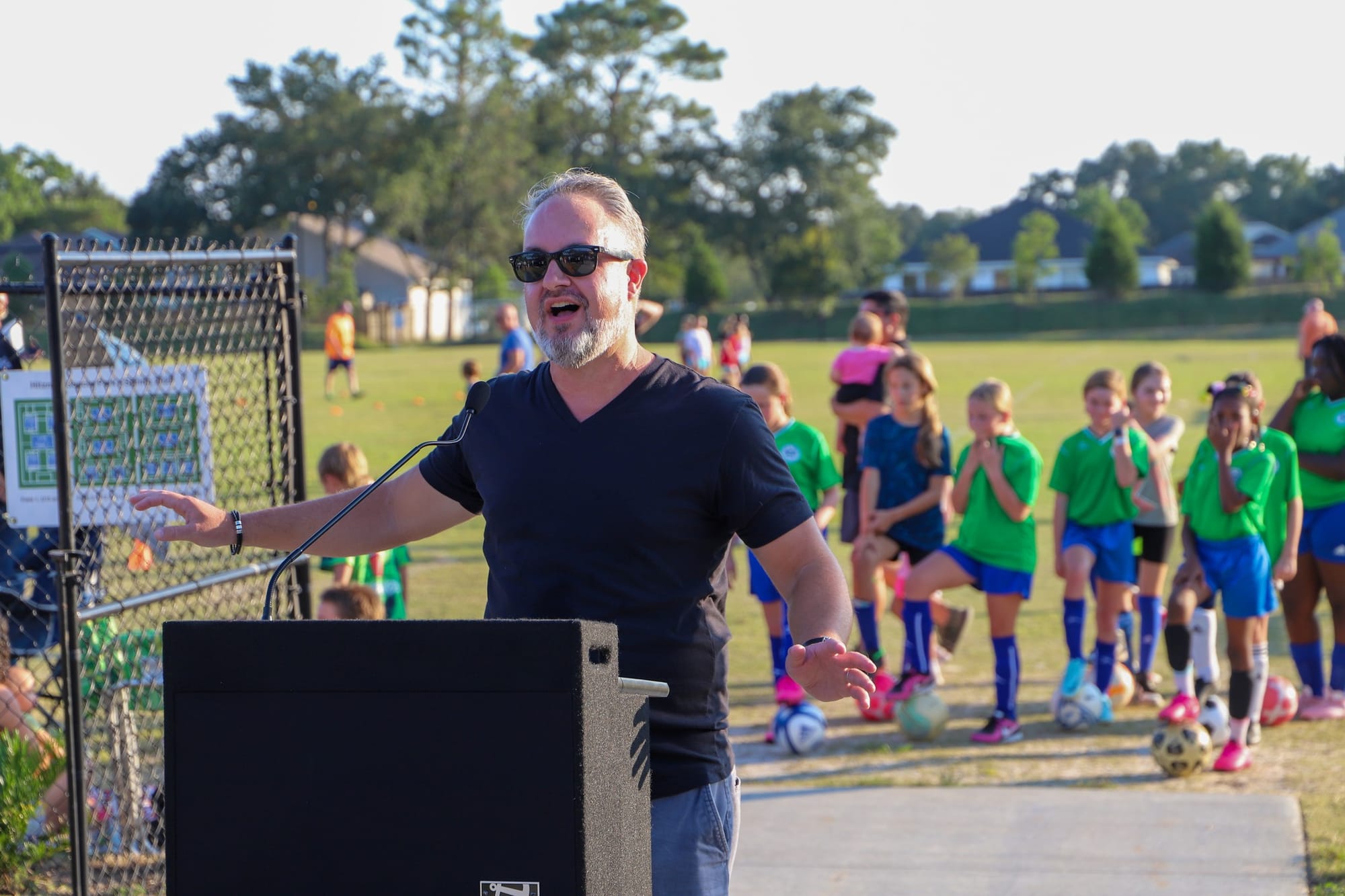 Phil Nickinson in a black T-shirt speaking in front of a group of children in soccer uniforms.